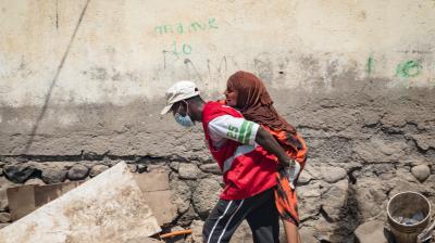 A person in a red vest and face mask carries another person on their back along a rough, debris-strewn street next to a worn wall.