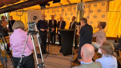 A group of people in formal attire stand and speak at a podium during a press event; several photographers and seated attendees are present.
