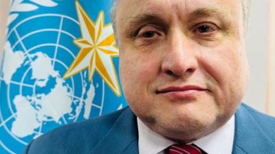 A man in a blue suit and red striped tie stands in front of a blue flag with a yellow star and a world map emblem.