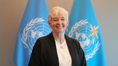 A person with short gray hair stands in front of two blue United Nations flags, wearing a black jacket over a white shirt and smiling at the camera.