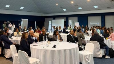 People seated at round tables in a large conference room, attentively listening to a presentation. The room is well-lit with white tablecloths and covered chairs.