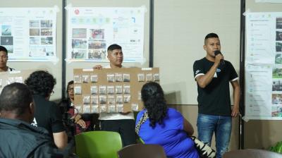 A man speaks into a microphone while another holds a display board with many small bags. Several people are seated, and posters with images and charts are on the wall behind them.