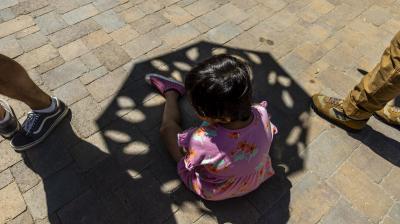 A child in a pink floral dress sits on a stone pavement, partially shaded by an umbrella, with three adults’ legs visible nearby.