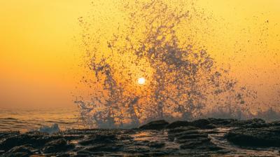 Ocean waves splash against rocks at sunset, with the sun visible through water droplets and an orange sky in the background.