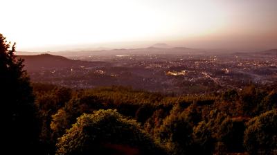 Elevated view of a city at sunset with dense trees in the foreground, city lights appearing as the sun sets, and mountains visible in the distant background.