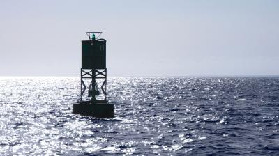 A green navigational buoy with a small light is floating in the open sea under bright sunlight and a clear sky.