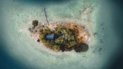 Aerial view of a small island with a single building, trees, a dock, and shallow water surrounding it.