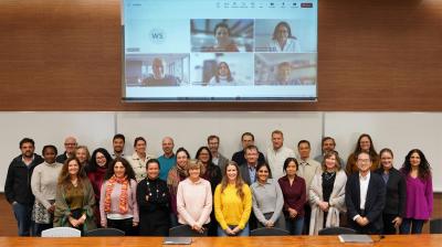 A group of people poses for a photo in a conference room, with a screen behind them displaying a video call with six participants.