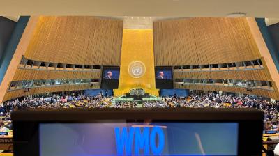 The United Nations General Assembly hall viewed from a desk labeled WMO, with a speaker visible on large screens above the podium.