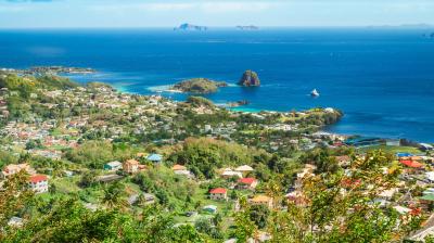 A high-angle view of a coastal town on a hillside overlooking a bay with islands and a yacht in the distance.