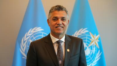 A man in a suit and tie standing in front of two United Nations flags.