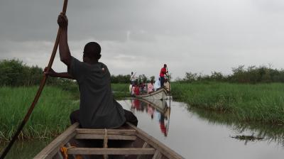 A person steers a wooden boat with a long pole through a grassy channel, following another boat filled with people.