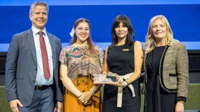 Four people stand together onstage, one holding an award plaque, all dressed in formal or semi-formal attire, posing for a group photo against a blue background.