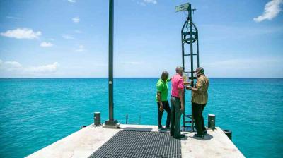 Three men stand on a pier next to a tall metal ocean monitoring device, with clear blue water and sky in the background.