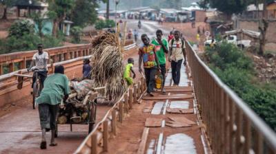 People walk and push carts across a narrow, rustic bridge in a rural area, with buildings and greenery visible in the background.