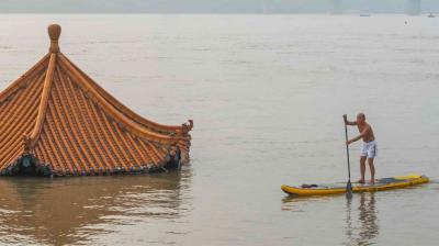 A person on a paddleboard navigates through floodwaters near a partially submerged traditional gabled roof.