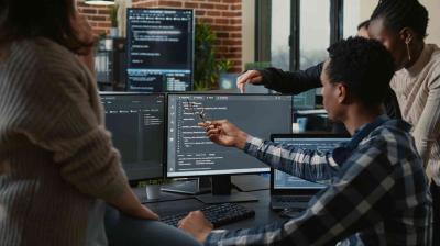 Group of people collaborating at a desk with multiple computer monitors displaying code in a modern office setting.
