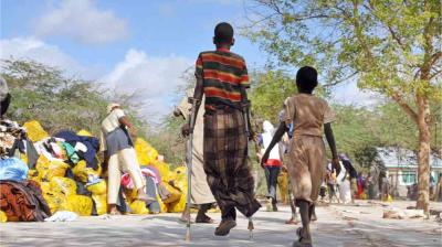 A child with crutches walks beside another child on a sunny street, with people sorting through yellow bags in the background.