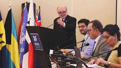 A man speaks at a podium during a conference, with four seated panelists and several international flags in the background.