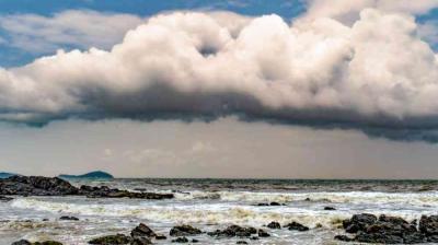 Large, dark storm clouds hang over a rocky shoreline with waves crashing onto the beach and a distant landmass on the horizon.