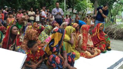 A group of women and children in colorful clothing sit outdoors, while people stand in the background, watching an event or gathering.
