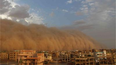 A massive sandstorm approaches a city with residential and commercial buildings under a partly cloudy sky.