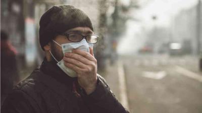 A person wearing glasses and a beanie holds a mask over their face while standing outside on a foggy or polluted street.