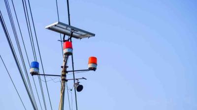 Utility pole with a solar panel, security camera, and red, blue, and orange warning lights against a clear blue sky.
