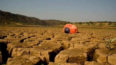 A person wearing a helmet is curled up on cracked, dry ground in a barren landscape with hills and sparse vegetation in the background.