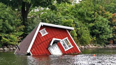 A red wooden house is partially submerged and tilted in a body of water, surrounded by trees and greenery.
