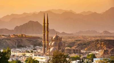 A mosque with two tall minarets stands in a city with desert mountains in the background during sunset.