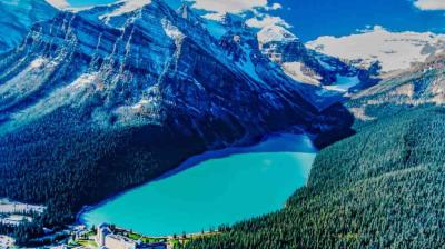 A turquoise lake surrounded by dense forest sits below tall, snow-capped mountains under a blue sky. Buildings are visible near the shoreline.