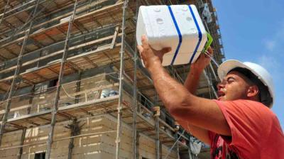 Construction worker in a hard hat lifts a white insulated block in front of a building covered in scaffolding under a blue sky.