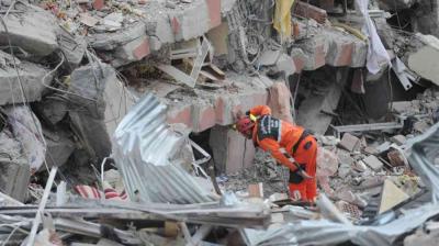 A rescue worker in orange gear searches through debris and collapsed buildings after a disaster, surrounded by rubble and broken structures.