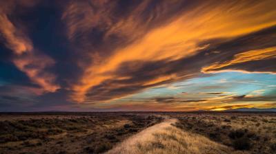 A dirt path winds through dry grass under a dramatic sunset sky with orange and blue clouds stretching across the horizon.