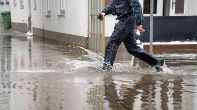 A person in dark clothing walks through a flooded street with water reaching above the ankles in a residential area.
