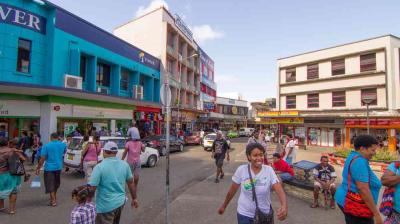 Busy street scene in a town with people walking, colorful buildings, parked cars, and storefronts on a sunny day.