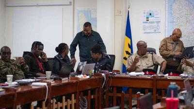 Several uniformed officers and officials sit and stand around a conference table with laptops and documents in a meeting room, with maps and a flag visible on the walls.