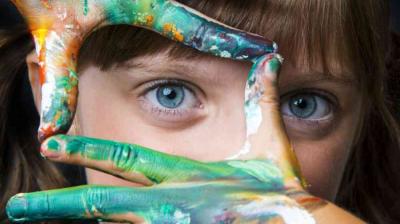 A child with blue eyes frames their face with hands covered in colorful paint, looking directly at the camera.