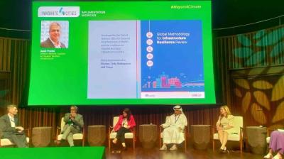 Five panelists sit on stage during a conference presentation, with a green backdrop displaying a project on global infrastructure resilience review.
