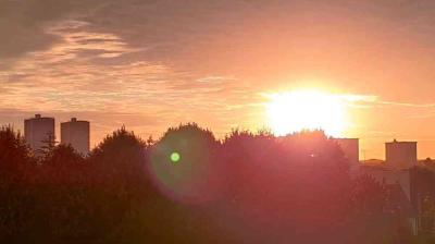 The sun rises over a city skyline with trees in the foreground and tall buildings silhouetted against an orange sky.