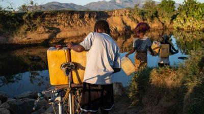 Three people collect water from a river, carrying yellow containers; one person stands beside a bicycle in the foreground. Dry terrain and mountains are visible in the background.