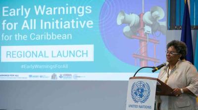 A woman speaks at a podium with the United Nations emblem during the Early Warnings for All Initiative regional launch event for the Caribbean. A presentation slide is displayed in the background.