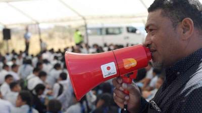 A man speaks into a red megaphone while a group of people sit on the ground under a large canopy in the background.