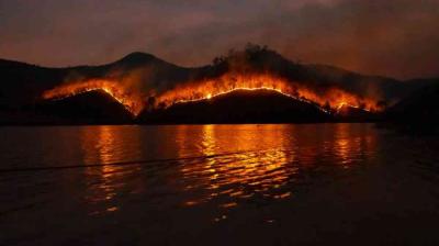 A wildfire burns along the ridge of a hill at night, with flames reflected in the water below.