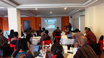 A group of people sit at round tables in a conference room, viewing a presentation projected on a screen at the front of the room.
