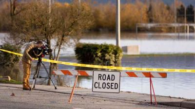 A person films a flooded street behind a "Road Closed" sign and caution tape; trees and water are visible in the background.