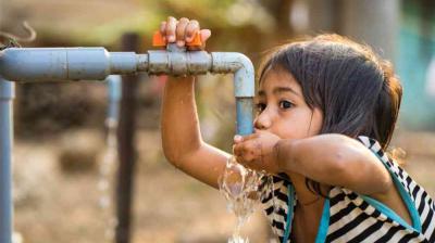 A young girl drinks water from a blue outdoor faucet, using one hand to hold the tap and the other to catch the water.