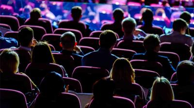 Audience members are seated in rows of theater-style chairs, facing a stage or screen, with dim lighting in the room.
