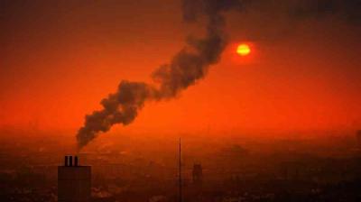 Industrial chimney emitting dark smoke into an orange sky at sunset, with the sun partially obscured by haze over a cityscape.